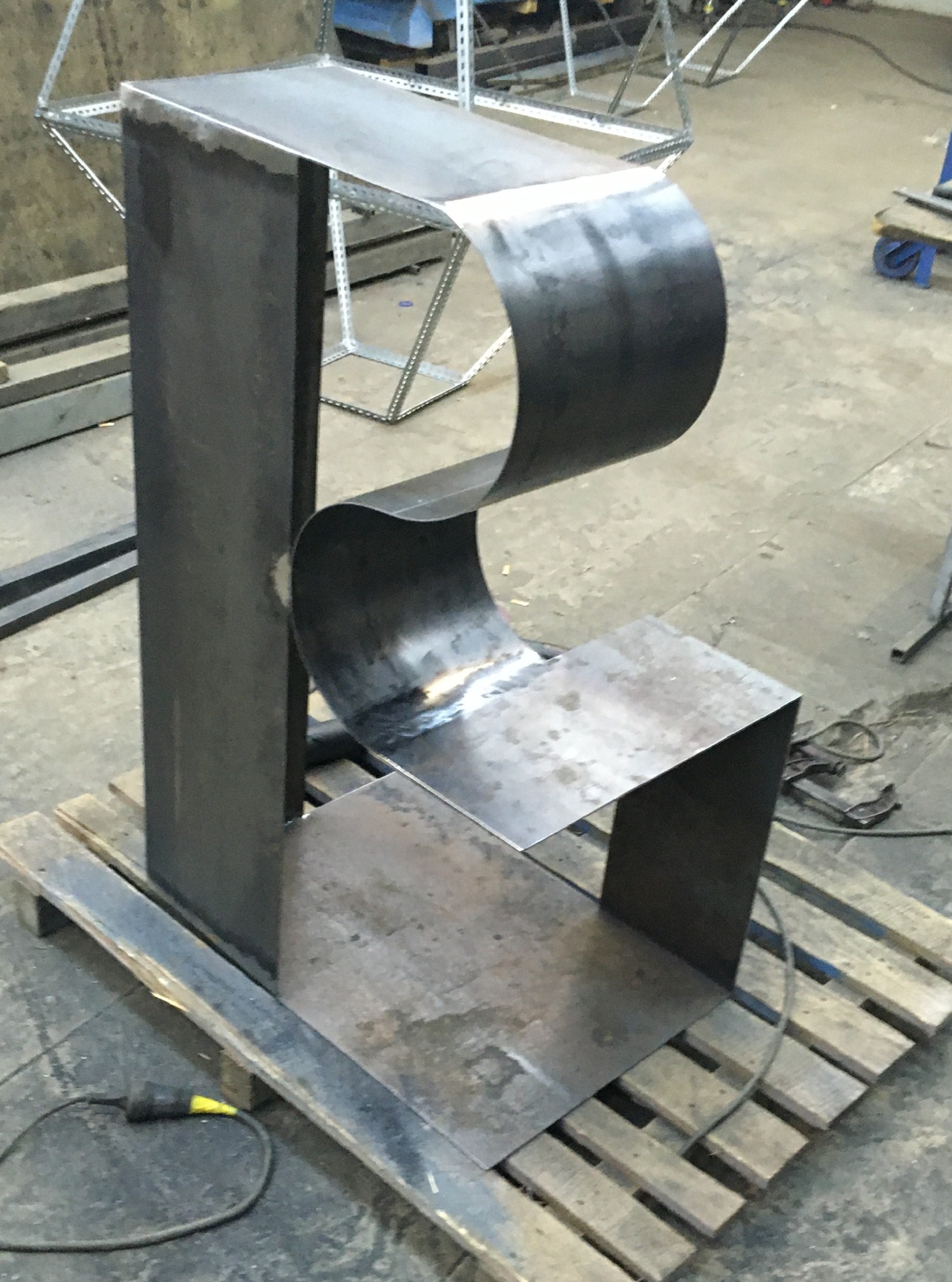 Surowy, spawany metalowy mebel o nietypowym kształcie, przypominającym literę E, na palecie w warsztacie. Widoczne ślady spawania i obróbki.