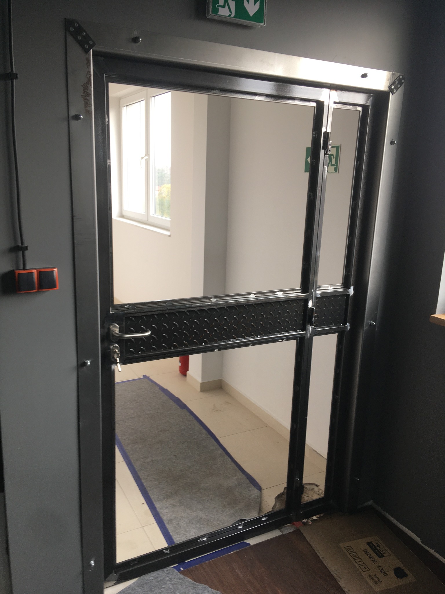 Metalowa, dwuskrzydłowa konstrukcja drzwiowa z szybami, zamontowana w szarej ścianie. Widoczny element ewakuacyjny, podłoga wyłożona płytkami.