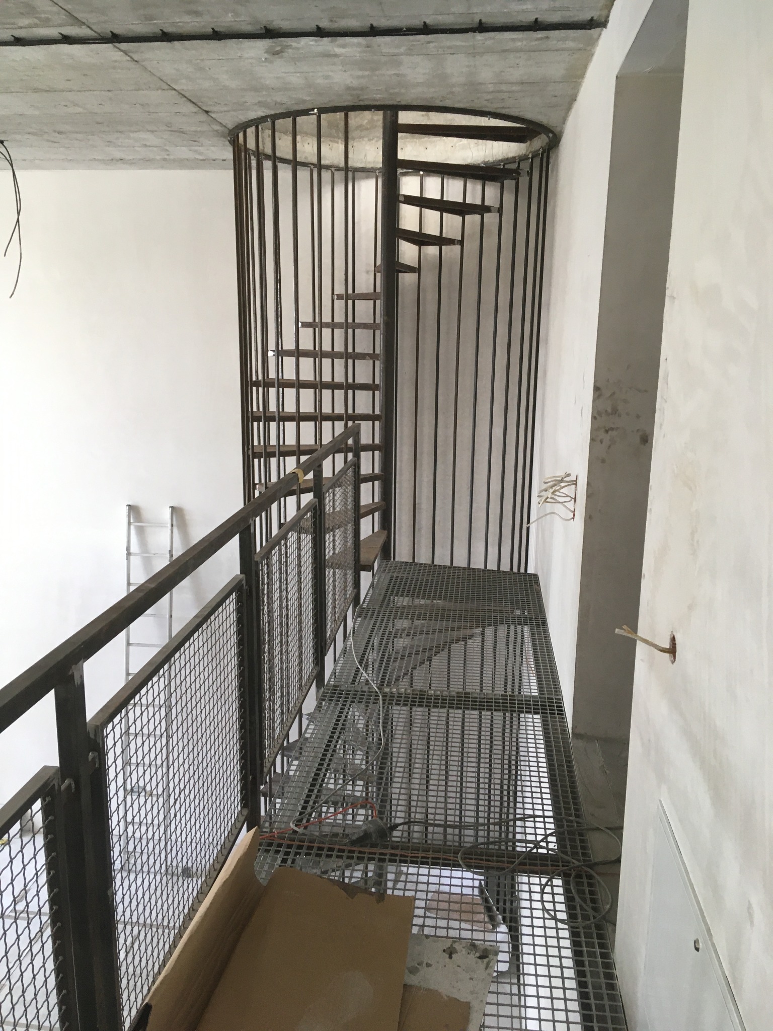 Metalowa konstrukcja schodów spiralnych z ażurową podłogą na antresoli w surowym wnętrzu. Widoczne niedokończone ściany i sufit.