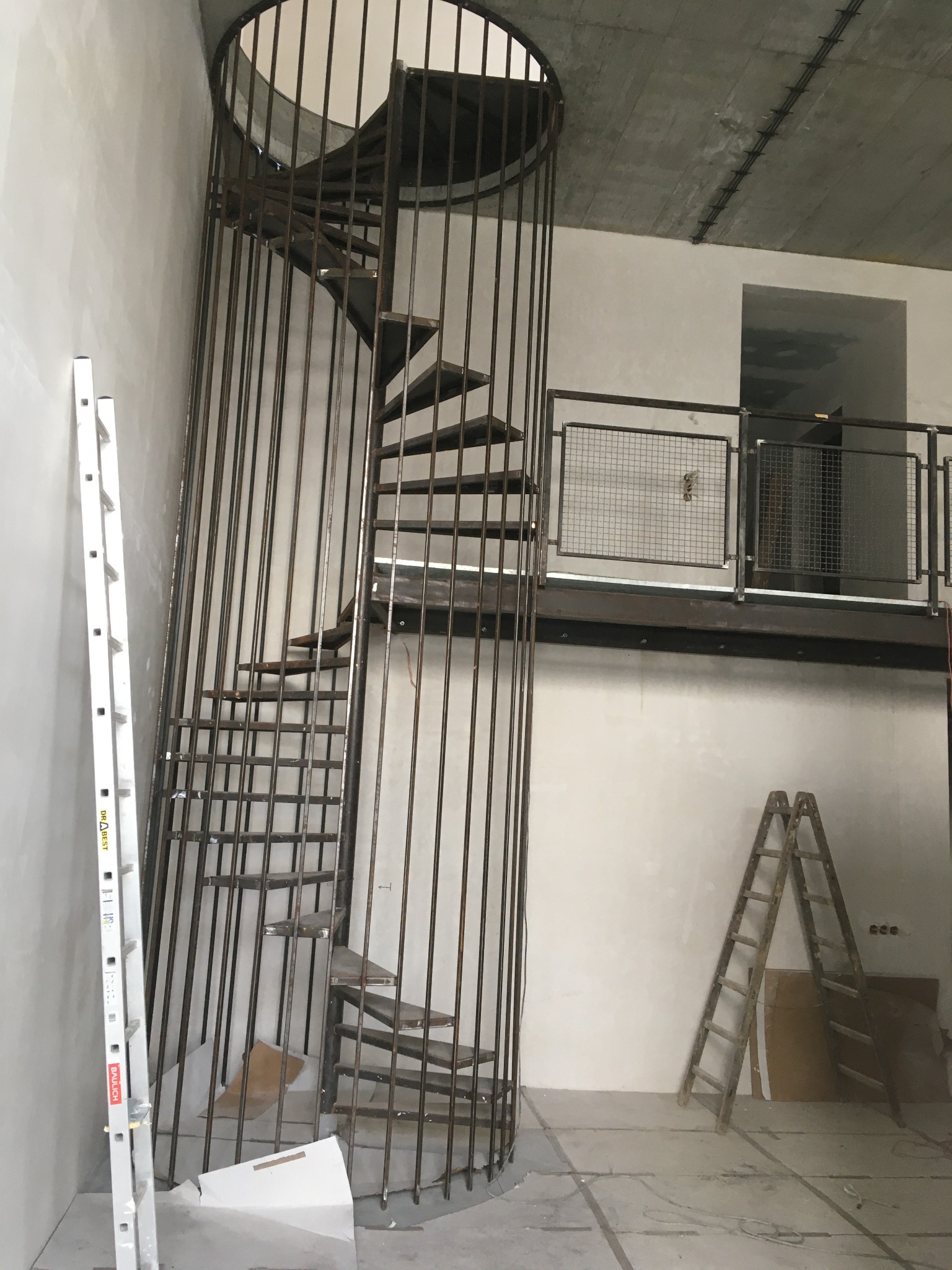 Metalowe schody spiralne w klatce z prętów, widoczne drabiny i balustrada na antresoli w surowym wnętrzu. Prace wykończeniowe w toku.