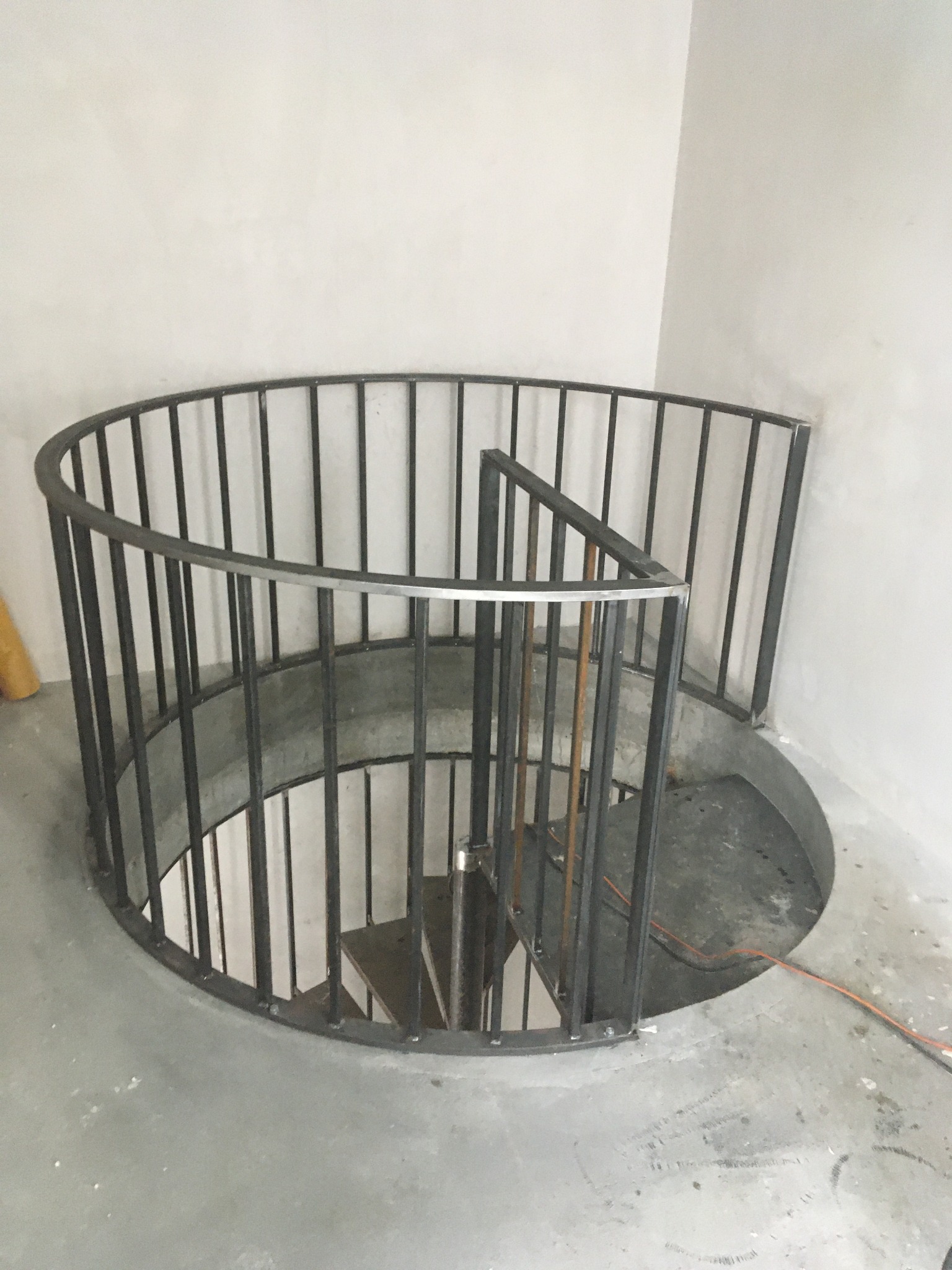 Spiralne schody metalowe z czarną balustradą w surowym wnętrzu. Widoczne spawy i niedokończone wykończenie podłogi.