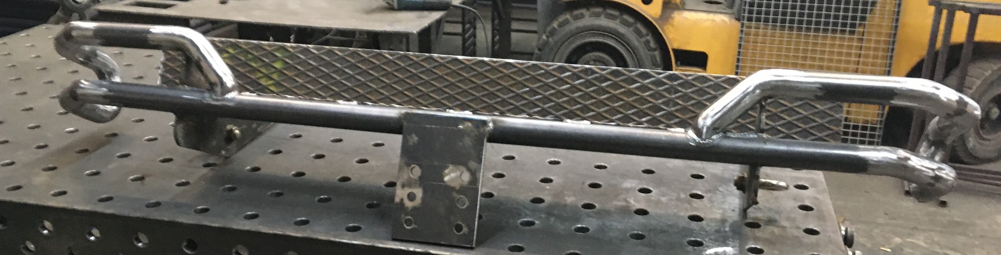 Metalowa konstrukcja z rur i blachy perforowanej na stole spawalniczym. Widoczne ślady spawania i obróbki. W tle koło maszyny budowlanej.