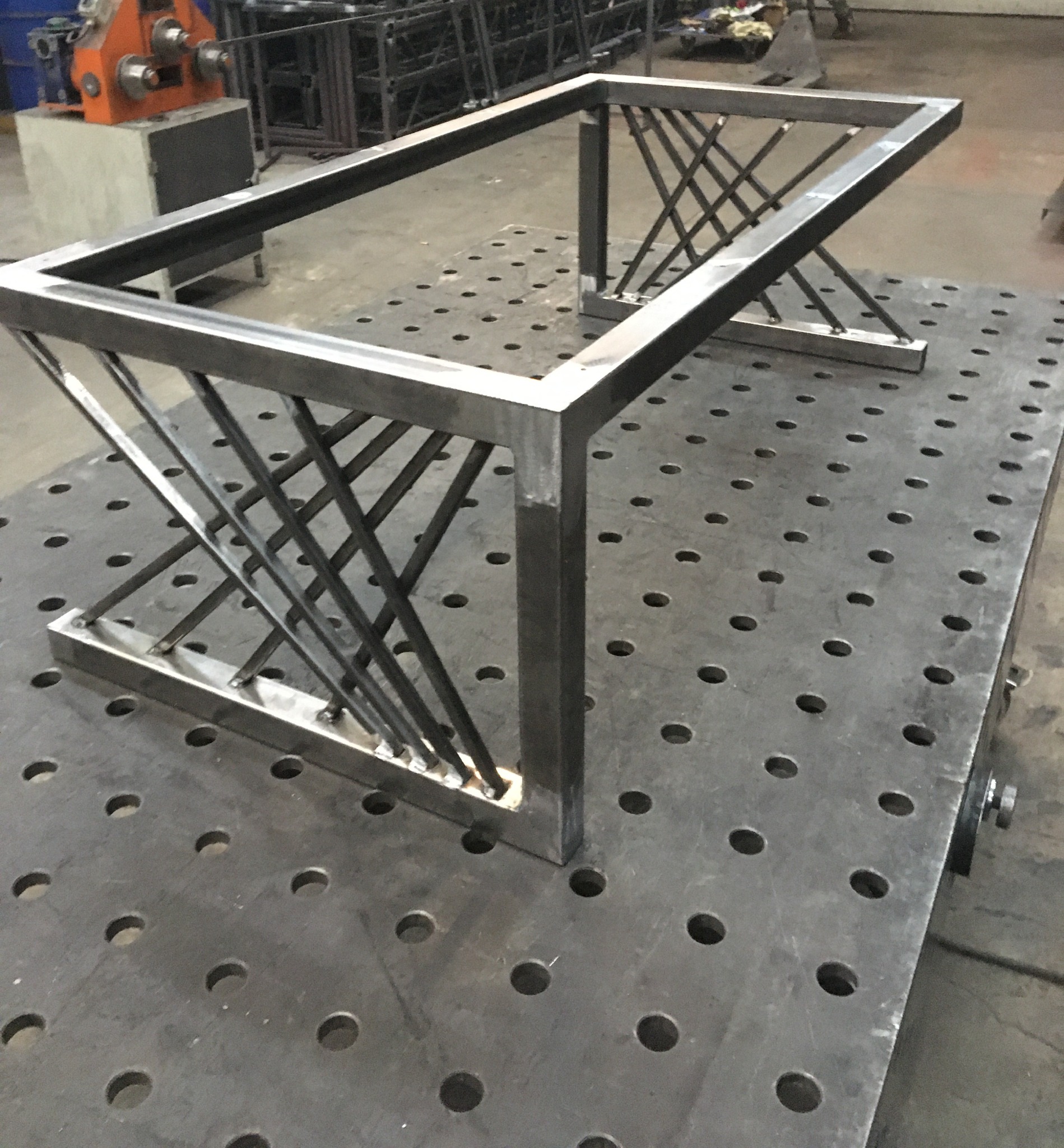 Stalowa konstrukcja ramy stołu z ukośnymi prętami, spawana na perforowanym stole spawalniczym. Widoczne ślady spawania i obróbki.