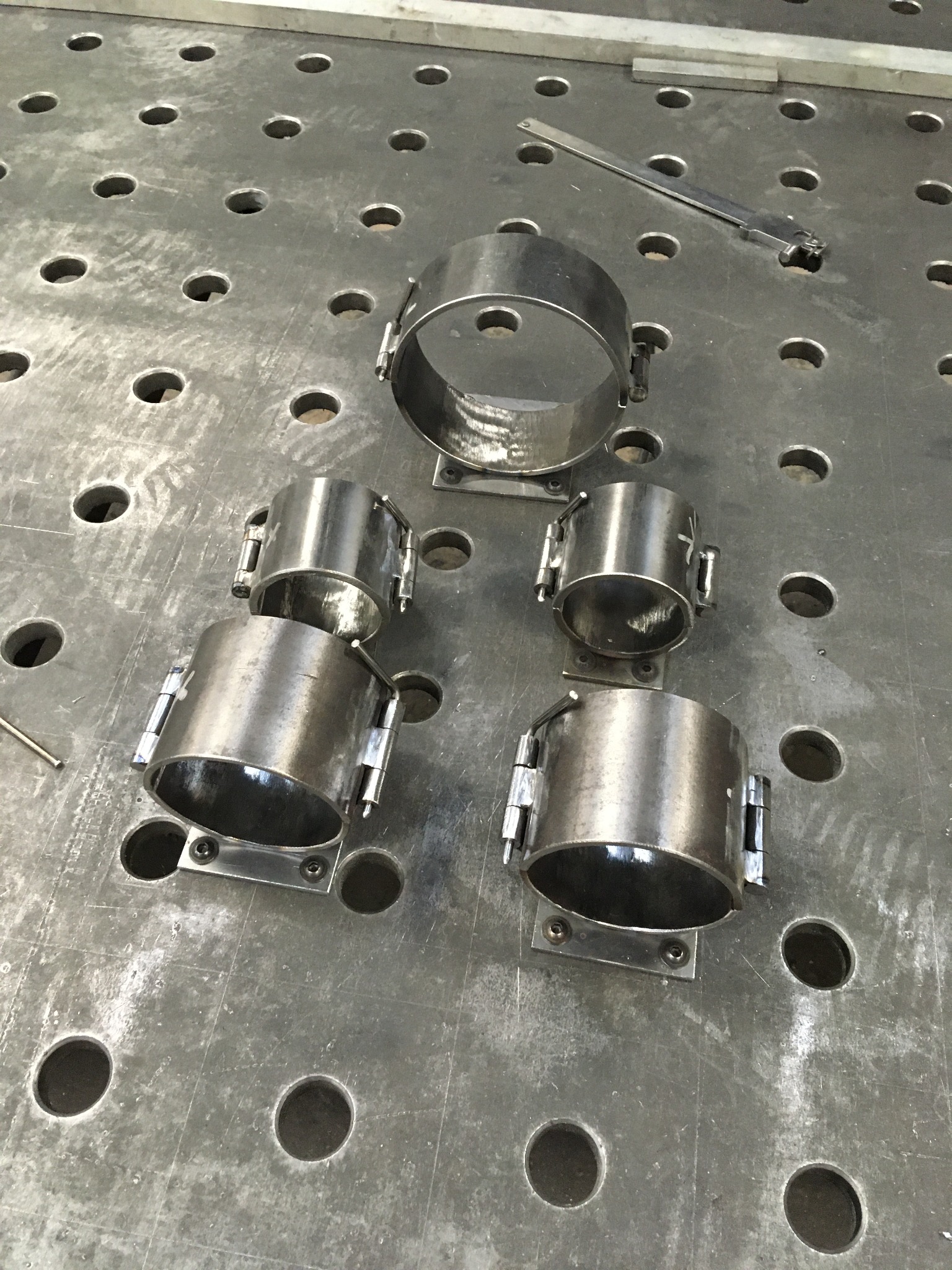 Metalowe obejmy z zamkami, wykonane w Warszawie, leżą na perforowanym stole warsztatowym. Widoczny suwmiarka. Precyzyjna obróbka metalu.