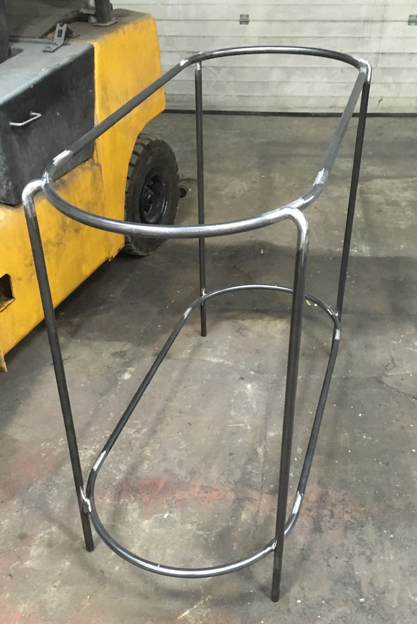 Surowa, metalowa konstrukcja z rurek o owalnym kształcie, spawana, w tle wózek widłowy. Widoczne spawy i niedoskonałości powierzchni.