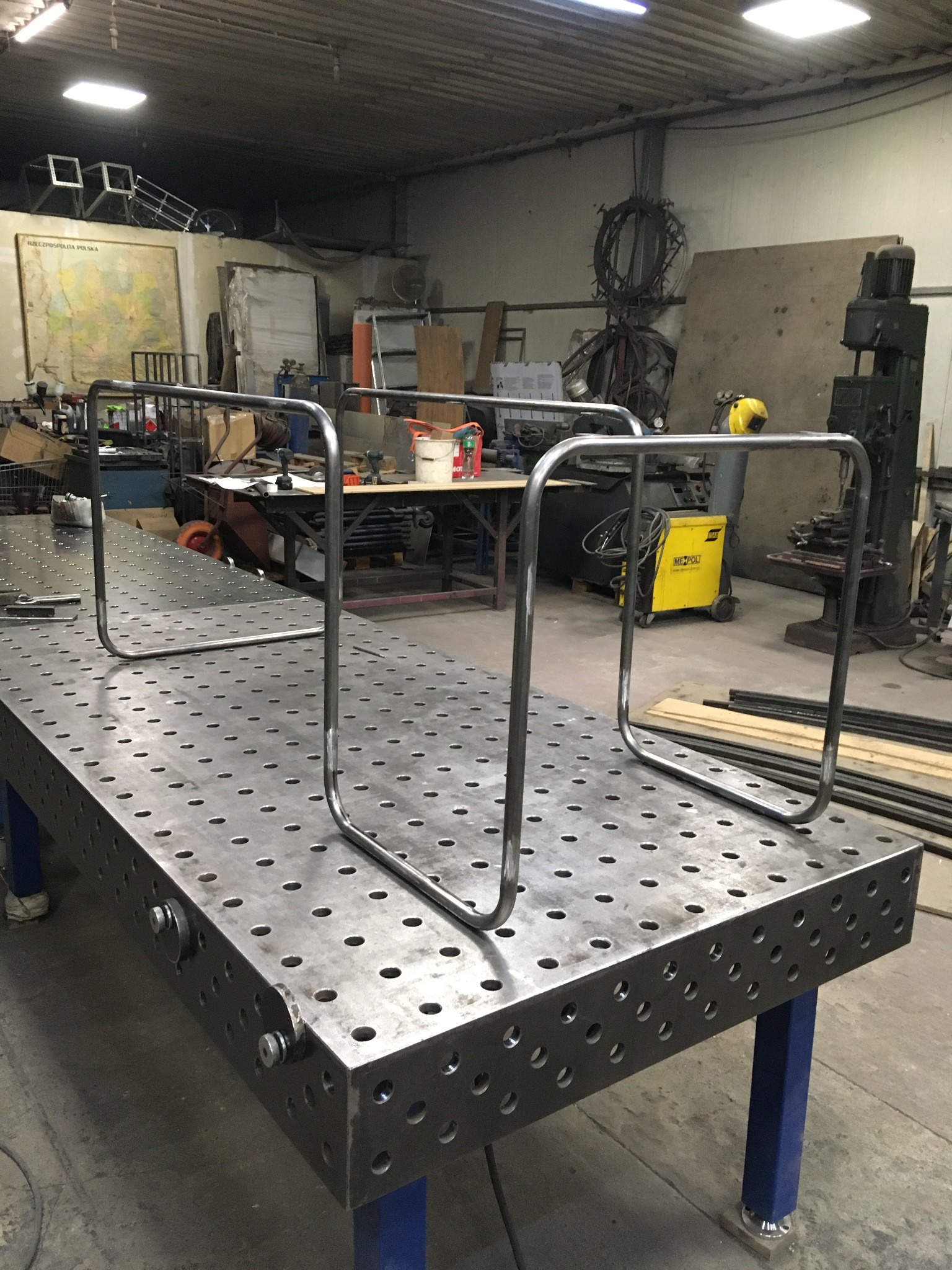 Metalowa poręcz w trakcie tworzenia na perforowanym stole spawalniczym. Widoczny warsztat z narzędziami i żółta spawarka. Surowe wykończenie.