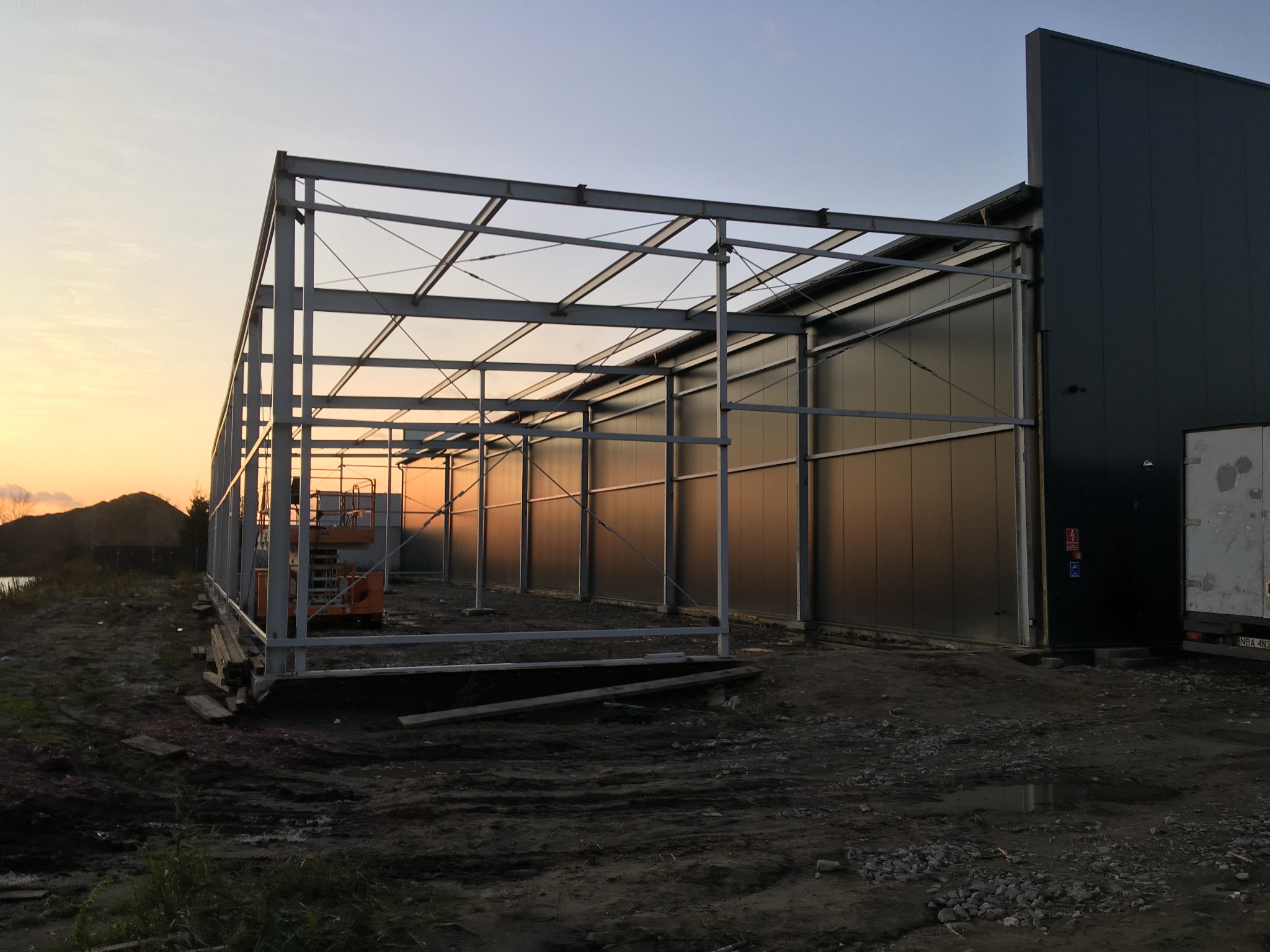 Konstrukcja stalowa hali w trakcie budowy o zmierzchu. Widoczny szkielet hali, ściany z blachy trapezowej i podnośnik koszowy wewnątrz konstrukcji.