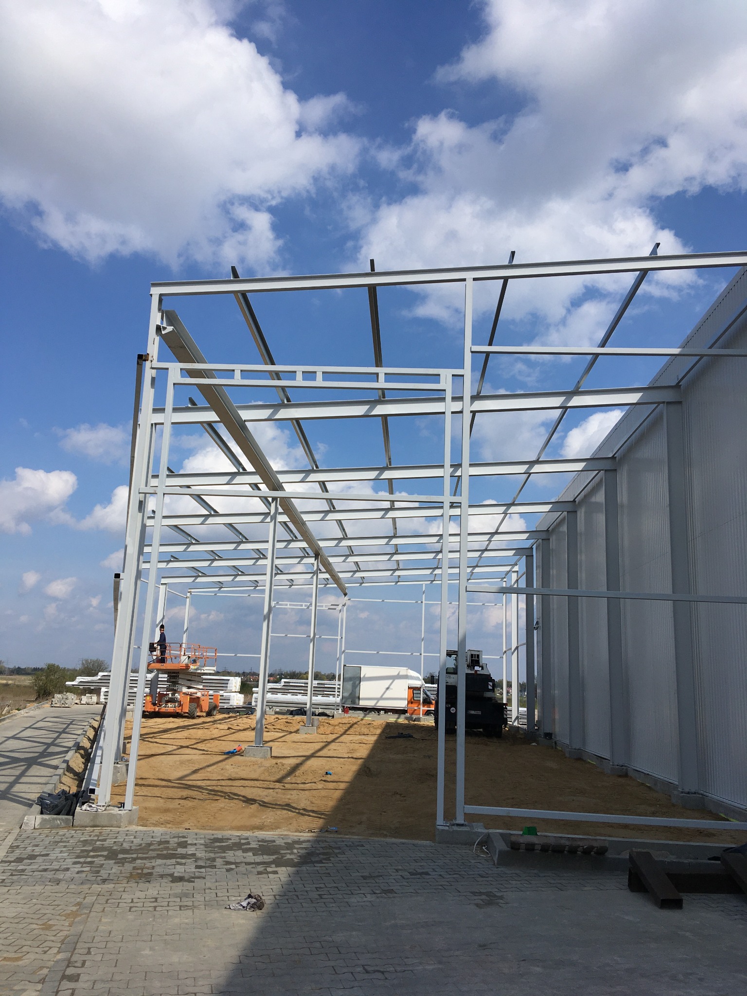 Wznoszenie szkieletu hali stalowej na tle błękitnego nieba. Widoczny podnośnik z pracownikiem, elementy konstrukcyjne i fragmenty obudowy hali. Jasne, naturalne światło.