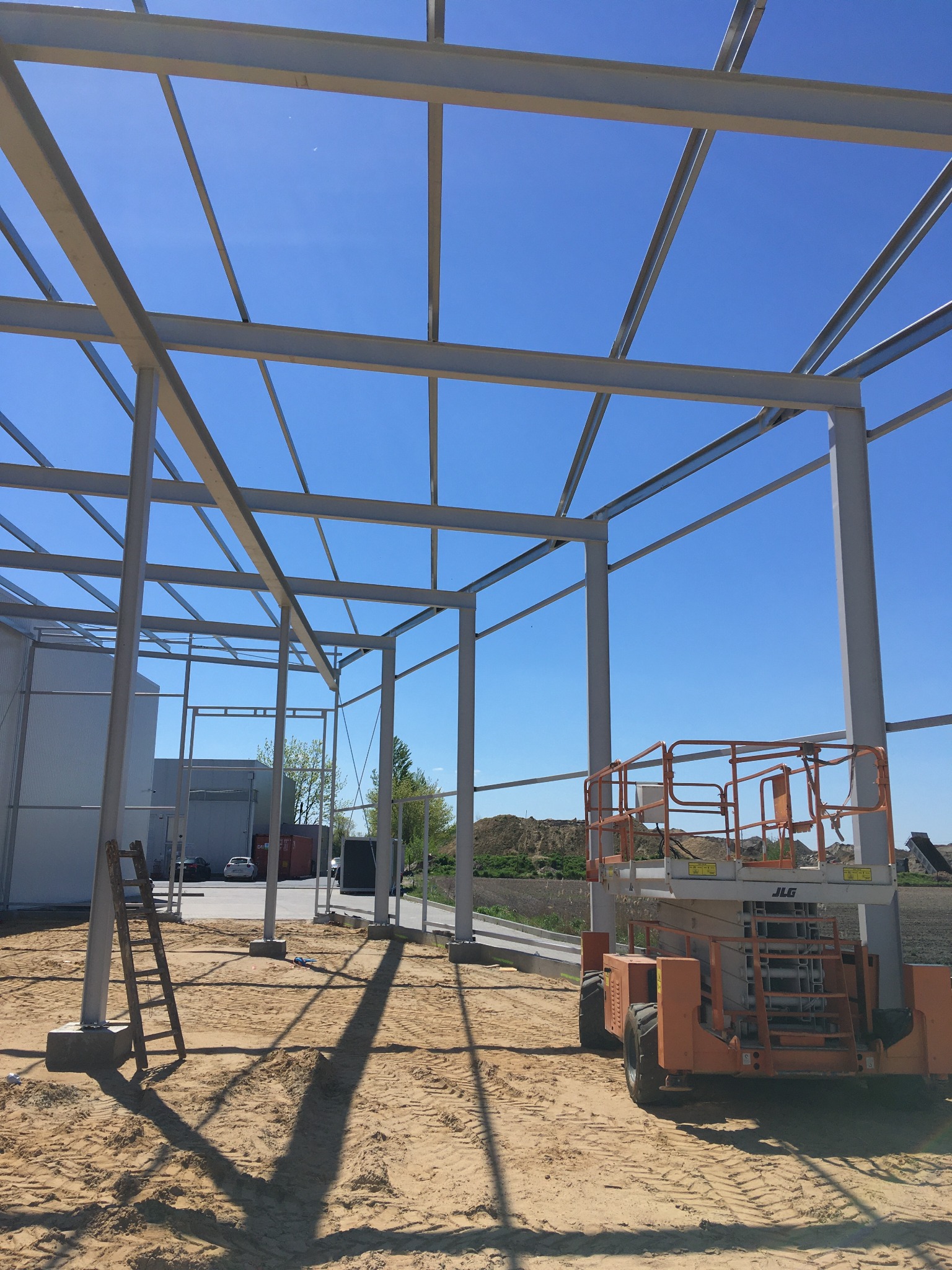 Stalowa konstrukcja hali w trakcie budowy, widoczna z dołu. Na pierwszym planie podnośnik i drabina. Błękitne niebo w tle. Elementy konstrukcji pomalowane na szaro.