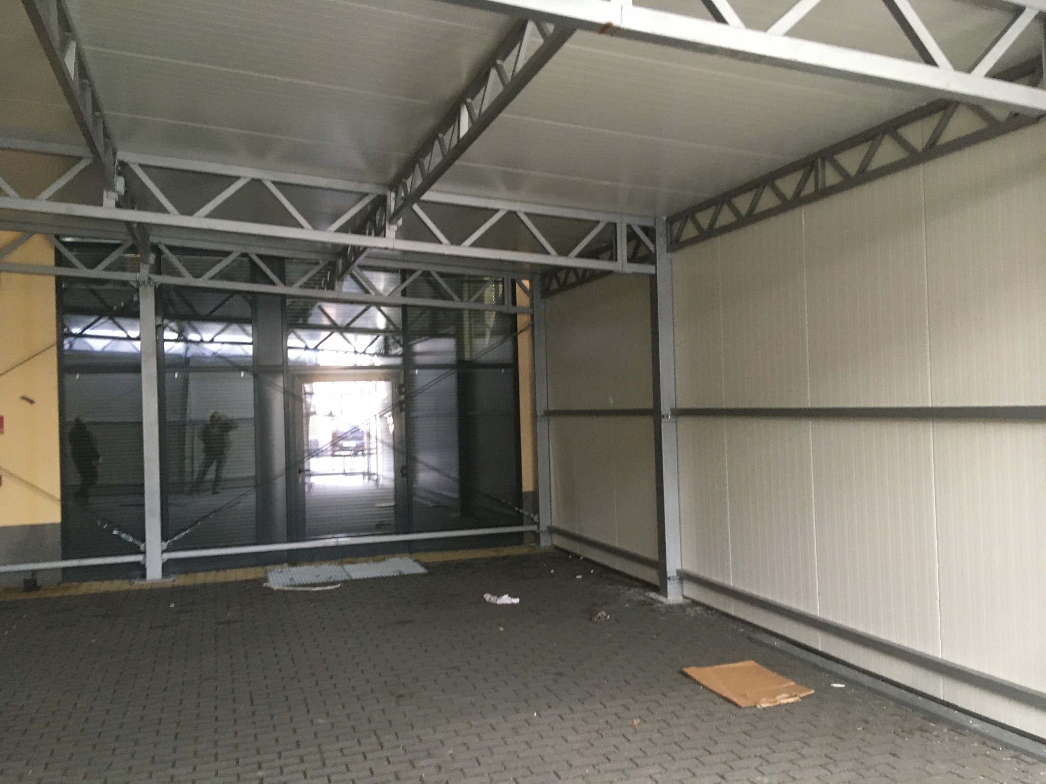 Wnętrze hali z metalową konstrukcją dachu i ścianami z paneli. Widoczne metalowe słupy i brama wjazdowa. Posadzka z kostki brukowej.