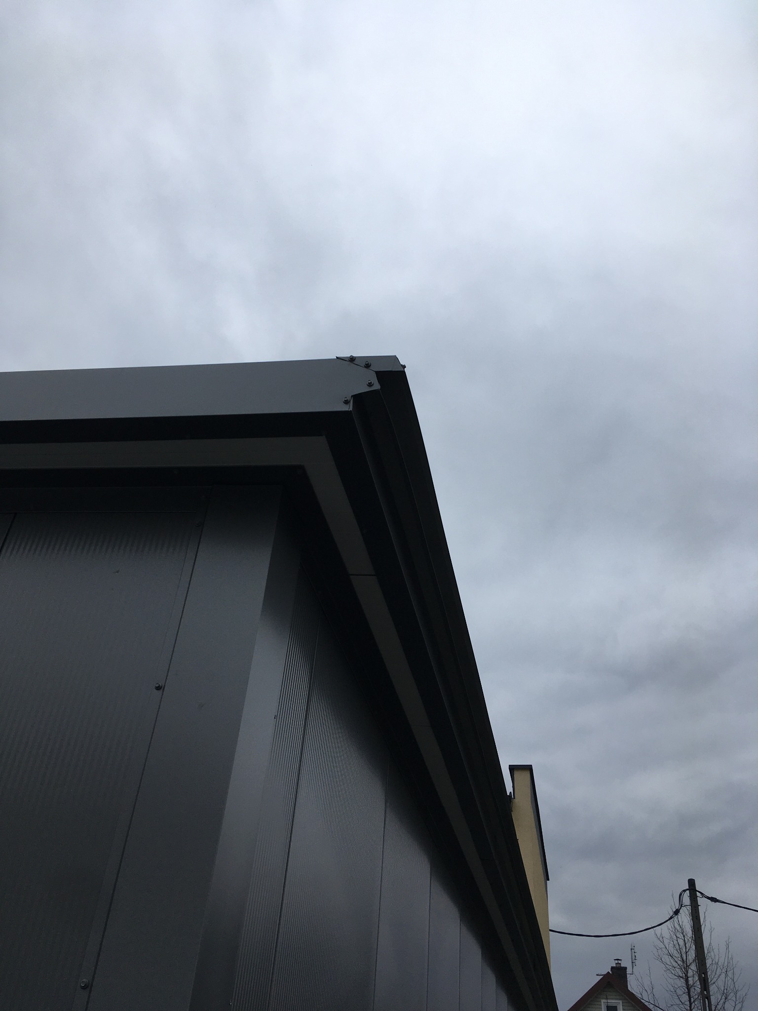 Szara hala z blachy trapezowej, widok z dołu na róg budynku i pochmurne niebo. Widoczne detale łączeń blach i fragment dachu sąsiedniego budynku.