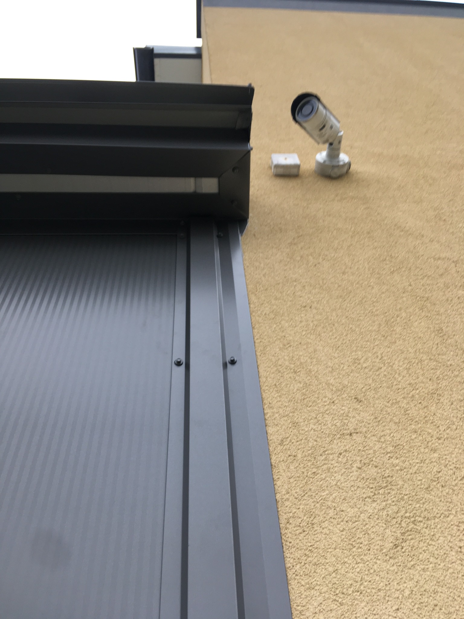 Szara, metalowa konstrukcja z widocznymi śrubami na tle beżowej ściany z kamerą przemysłową. Ujęcie z dołu, podkreślające solidność wykonania.