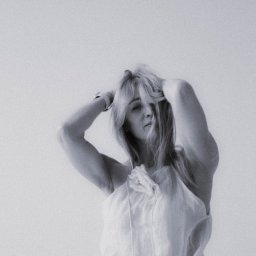 EWA POTOCZNA ConcEPt - Monochromatyczne, portretowe ujęcie kobiety z blond włosami, w zwiewnej bluzce, z rękami uniesionymi nad głową, na jednolitym, jasnym tle. Widoczne delikatne ziarno.