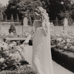 EWA POTOCZNA ConcEPt - Czarno-białe zdjęcie kobiety w koronkowej sukni, spacerującej wśród róż w ogrodzie, w słoneczny dzień. Długie włosy i zwiewny materiał sukni dodają elegancji.