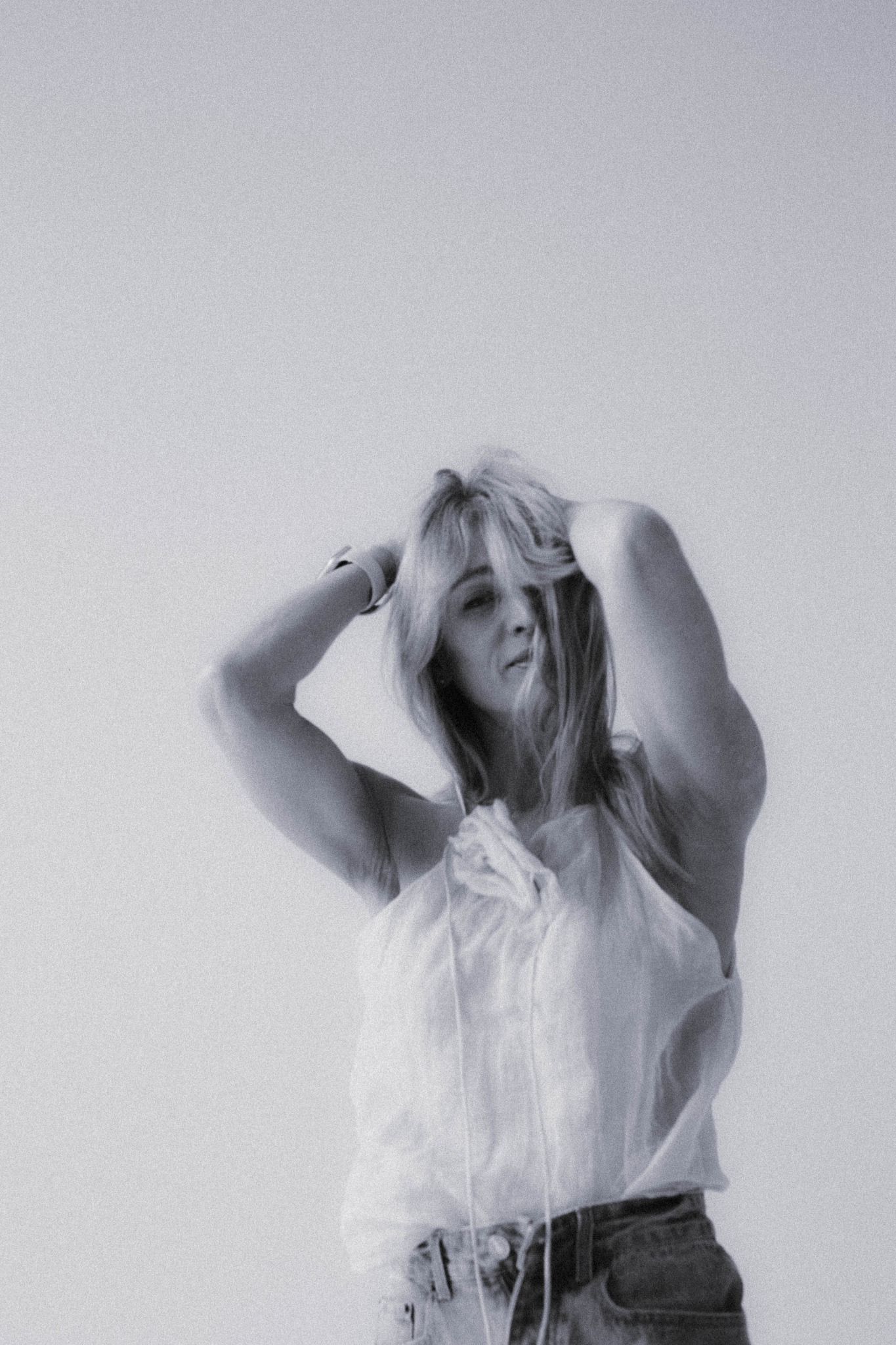 Monochromatyczne, portretowe ujęcie kobiety z blond włosami, w zwiewnej bluzce, z rękami uniesionymi nad głową, na jednolitym, jasnym tle. Widoczne delikatne ziarno.