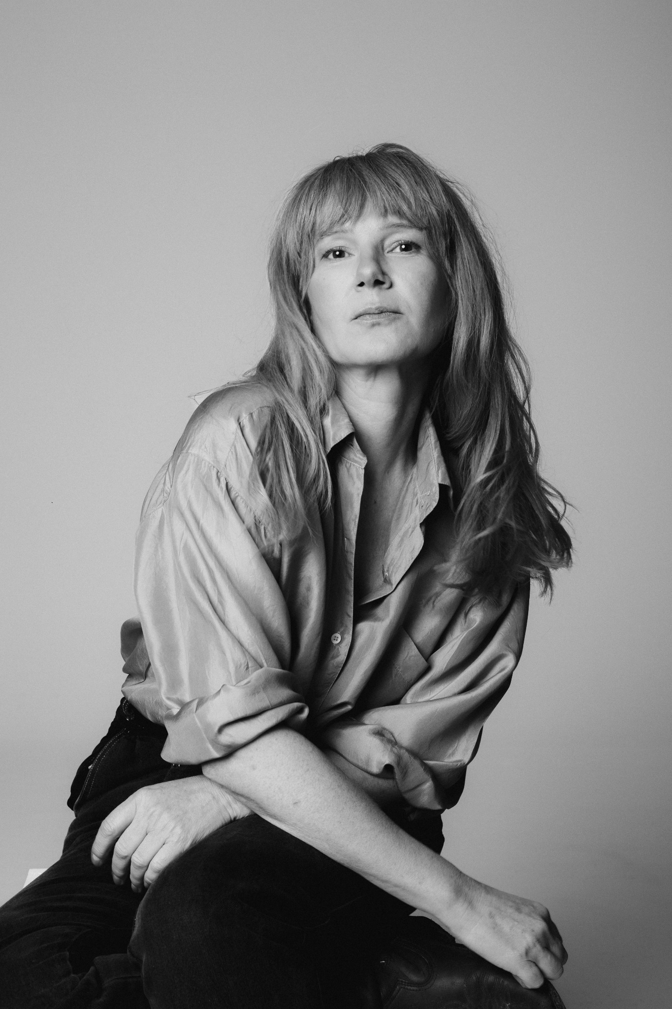 Czarno-białe, studyjne ujęcie kobiety z długimi włosami w satynowej koszuli. Skrzyżowane ręce, stonowane tło, naturalny wyraz twarzy.