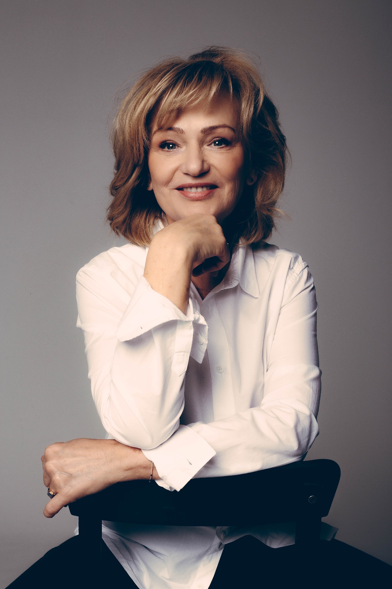 Portret kobiety w białej koszuli, opierającej się o krzesło. Delikatny uśmiech, naturalny makijaż, ciepłe oświetlenie. Profesjonalna sesja biznesowa.