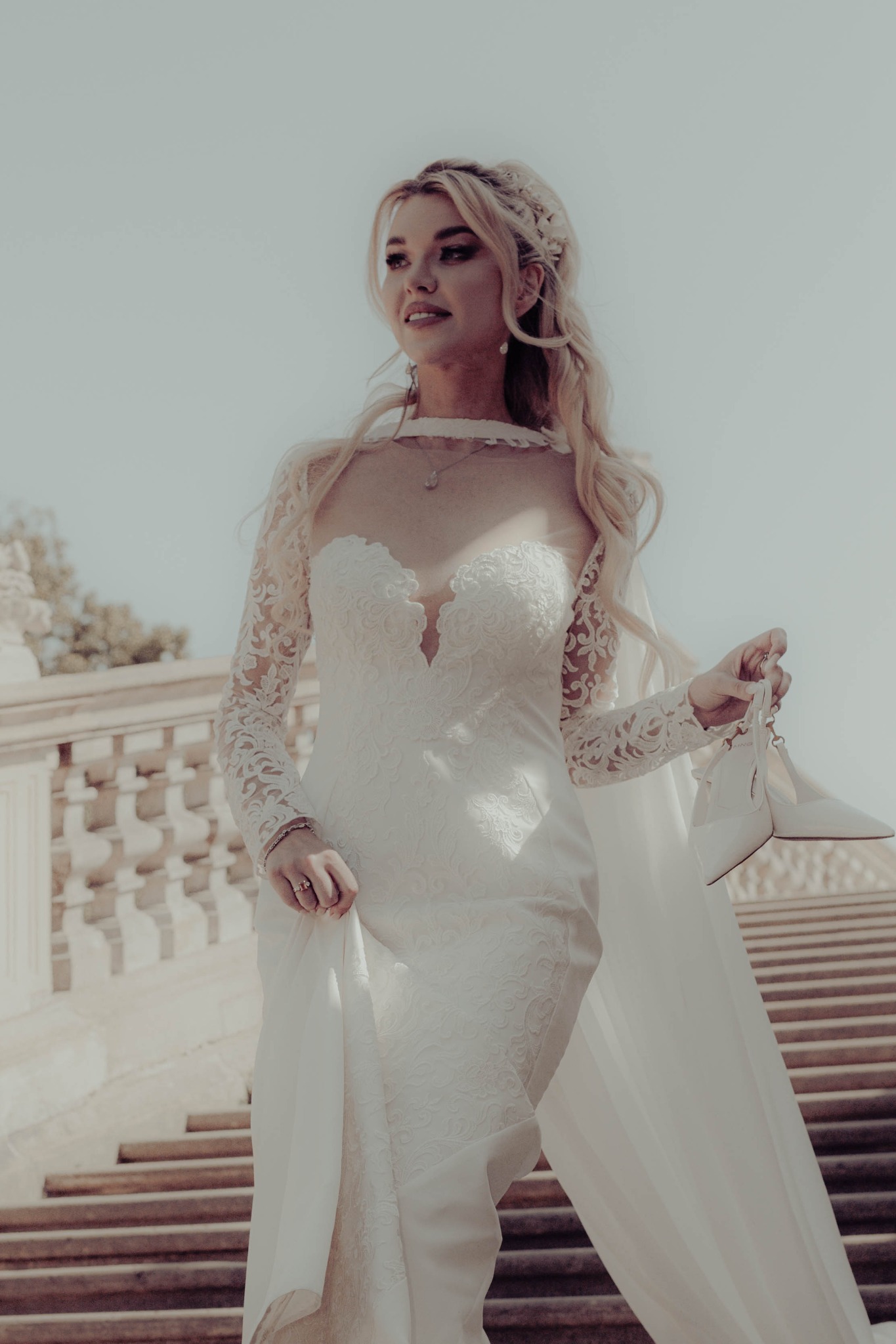 Panna młoda w koronkowej sukni z długim rękawem, trzymająca białe buty na schodach w słoneczny dzień. Upięte blond włosy z ozdobą.