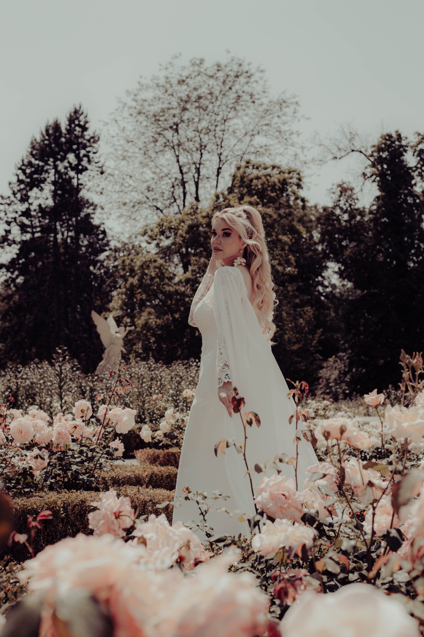 Elegancka blondynka w białej sukni w ogrodzie różanym, z posągiem anioła w tle. Romantyczna sceneria i delikatne światło tworzą bajkowy klimat.