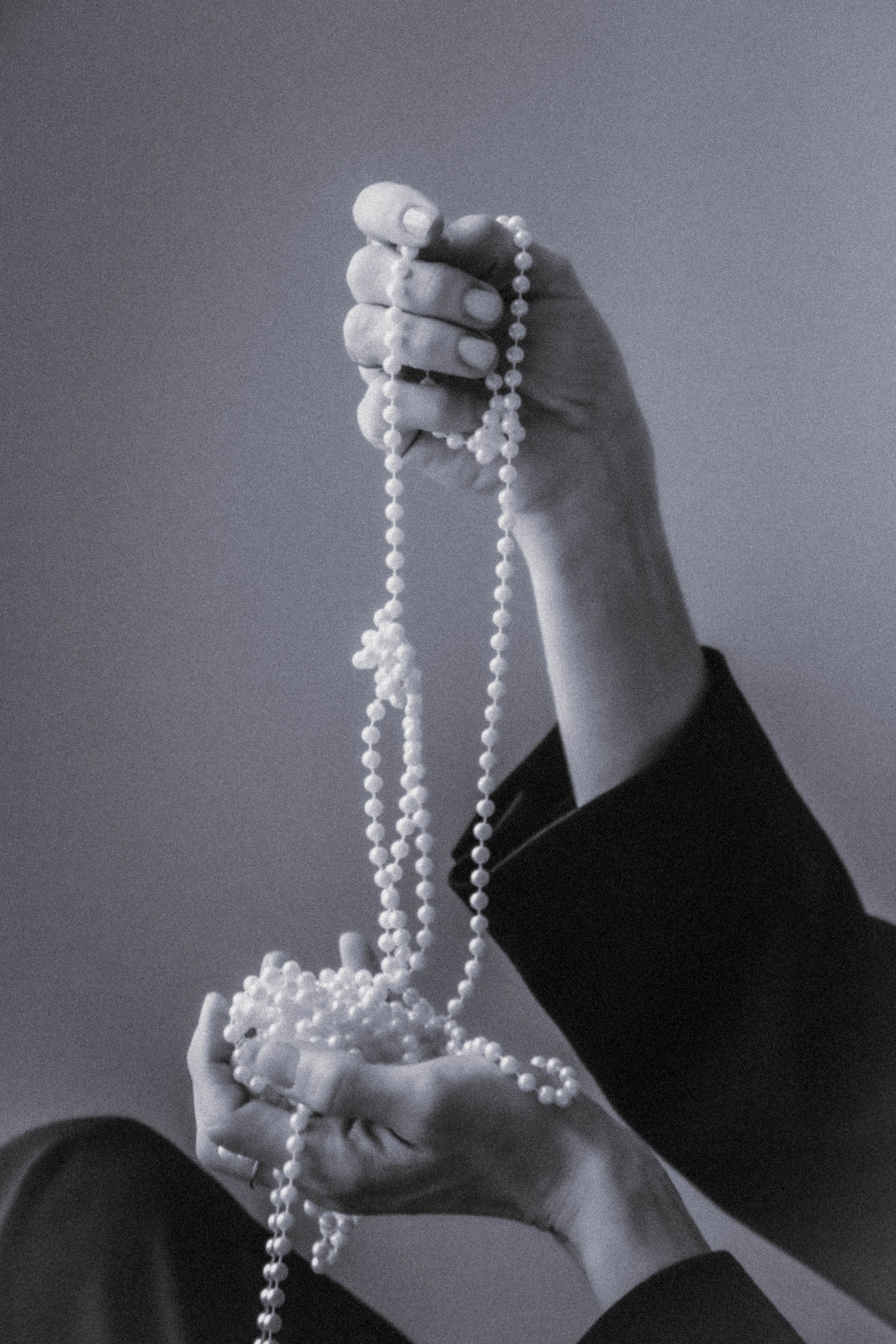 Dłonie kobiety w czarnym żakiecie trzymają sznur pereł na rozmytym, szarym tle. Elegancki minimalizm, skupienie na detalu i fakturze.
