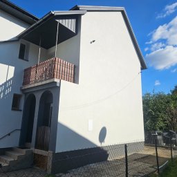 J.G. Future Company - Biały dom z balkonem i szarą podmurówką, widoczny zza ogrodzenia. Świeżo pomalowana elewacja w słoneczny dzień. Widoczne okna, drzwi wejściowe i fragment nieba.