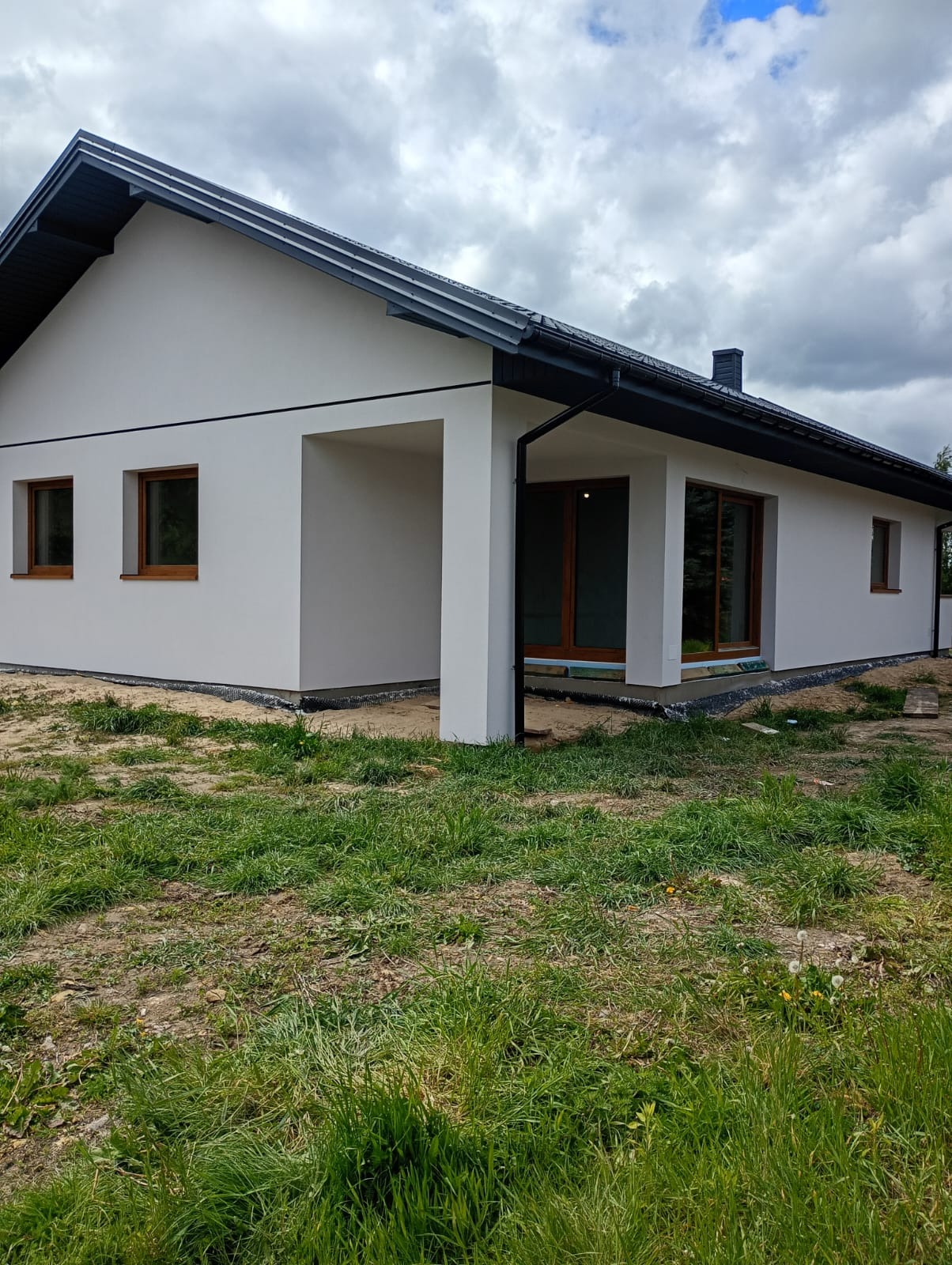 Nowoczesny, parterowy dom z białymi ścianami i ciemnym dachem, widoczny z poziomu trawnika. Okna z drewnianą ramą. Minimalistyczny styl, pochmurne niebo w tle.
