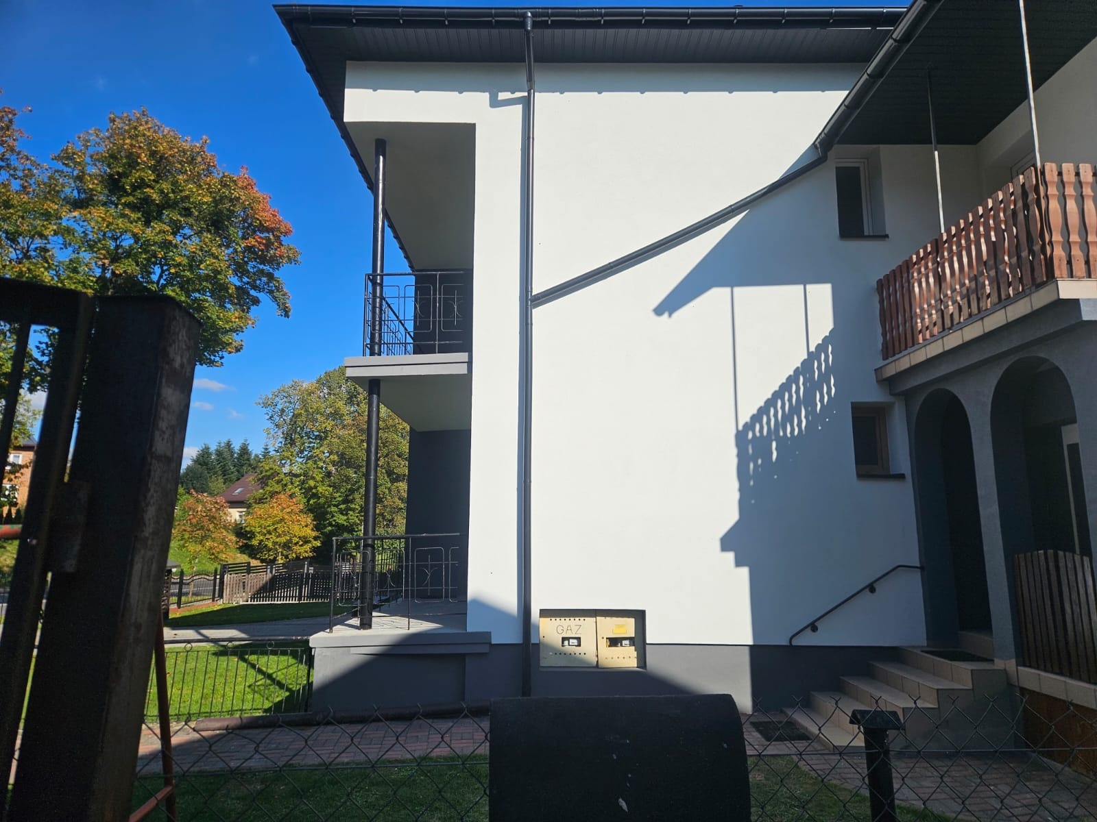 Biała elewacja budynku z balkonami i elementami w kolorze szarym, widoczna skrzynka gazowa, drzewa w tle i fragment ogrodzenia.