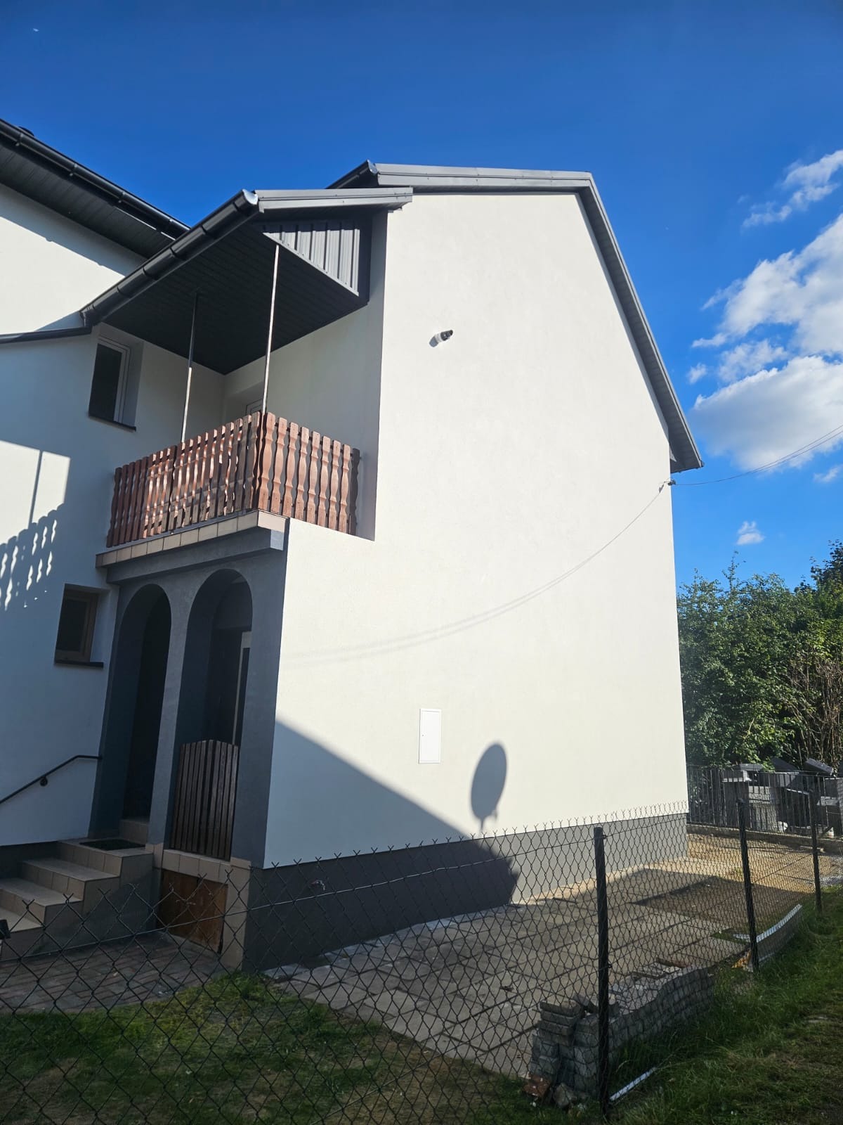 Biały dom z balkonem i szarą podmurówką, widoczny zza ogrodzenia. Świeżo pomalowana elewacja w słoneczny dzień. Widoczne okna, drzwi wejściowe i fragment nieba.