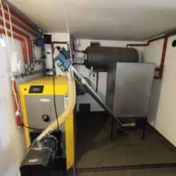 Żółto-szary piec CO marki LUMO z podajnikiem OKIPI w kotłowni. Widoczne rury instalacji grzewczej, zasobnik i elementy montażowe. Ujęcie z perspektywy podłogi.