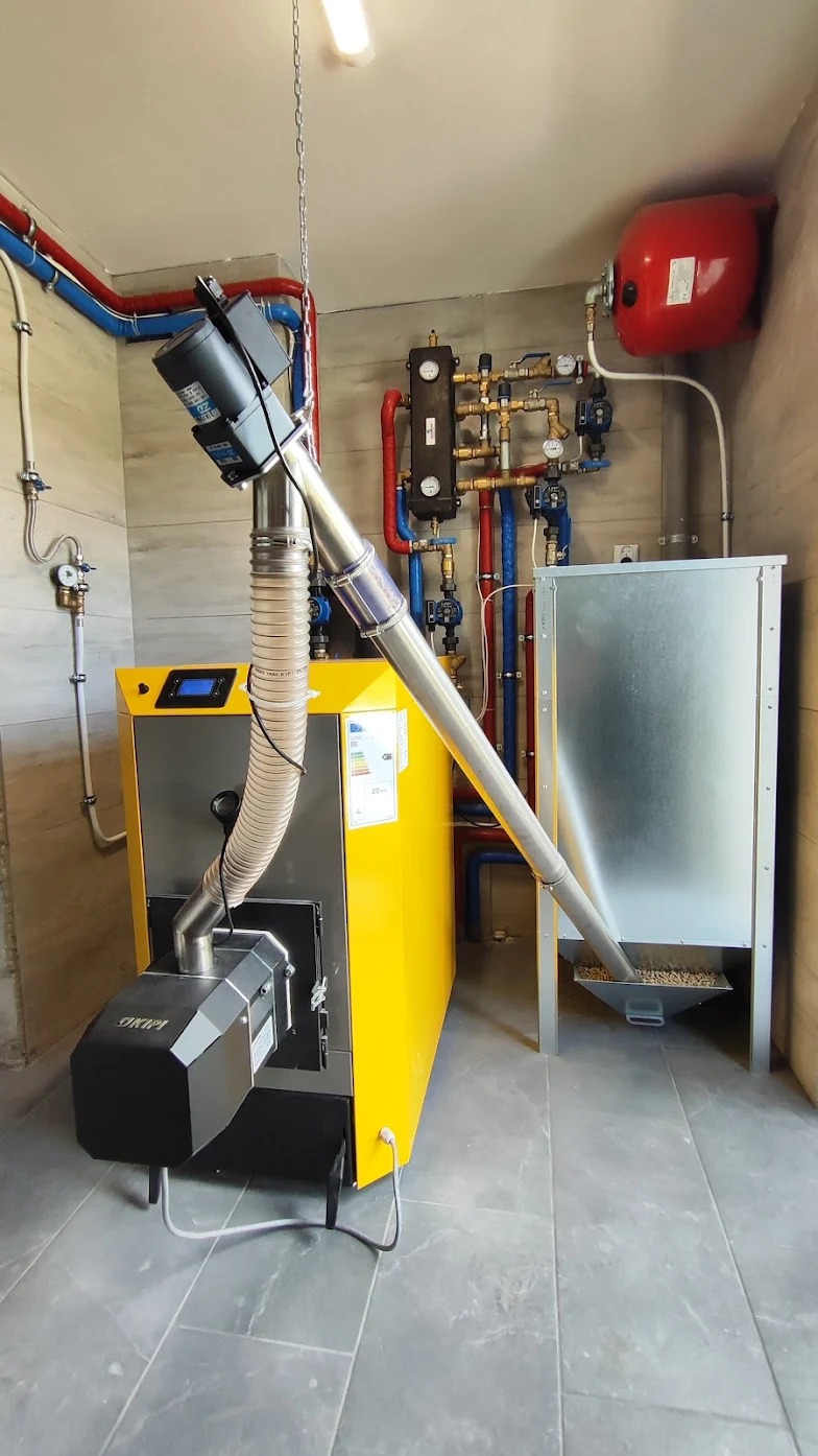 Żółty piec CO na pelet z podajnikiem, instalacja grzewcza z rurami miedzianymi i zbiornikiem wyrównawczym w kotłowni z szarą glazurą.