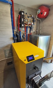 Żółty piec CO z czarnym panelem sterowania, instalacja z rur miedzianych i zaworów na ścianie wyłożonej płytkami imitującymi drewno, zbiornik wyrównawczy.