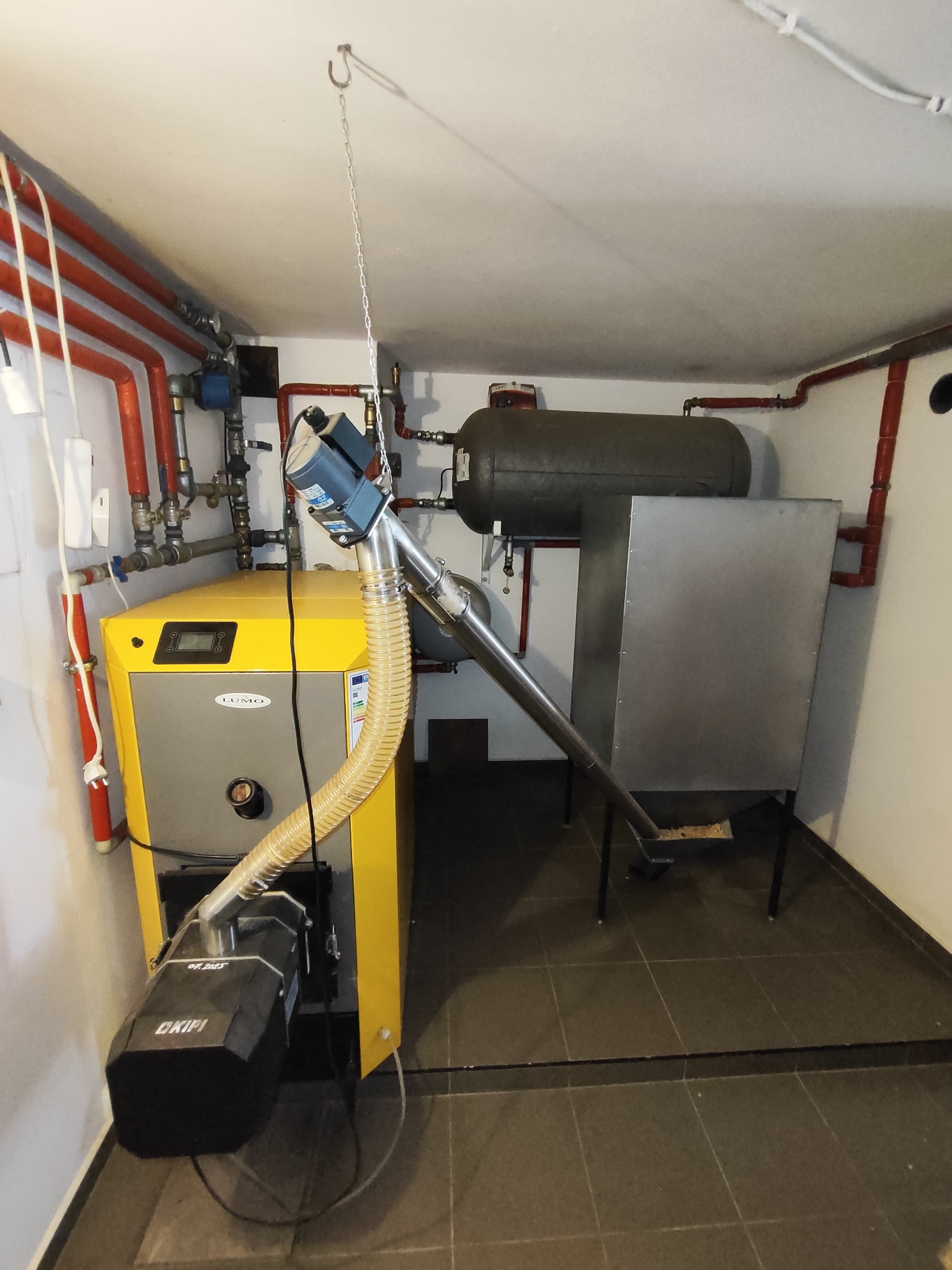 Żółto-szary piec CO marki LUMO z podajnikiem OKIPI w kotłowni. Widoczne rury instalacji grzewczej, zasobnik i elementy montażowe. Ujęcie z perspektywy podłogi.