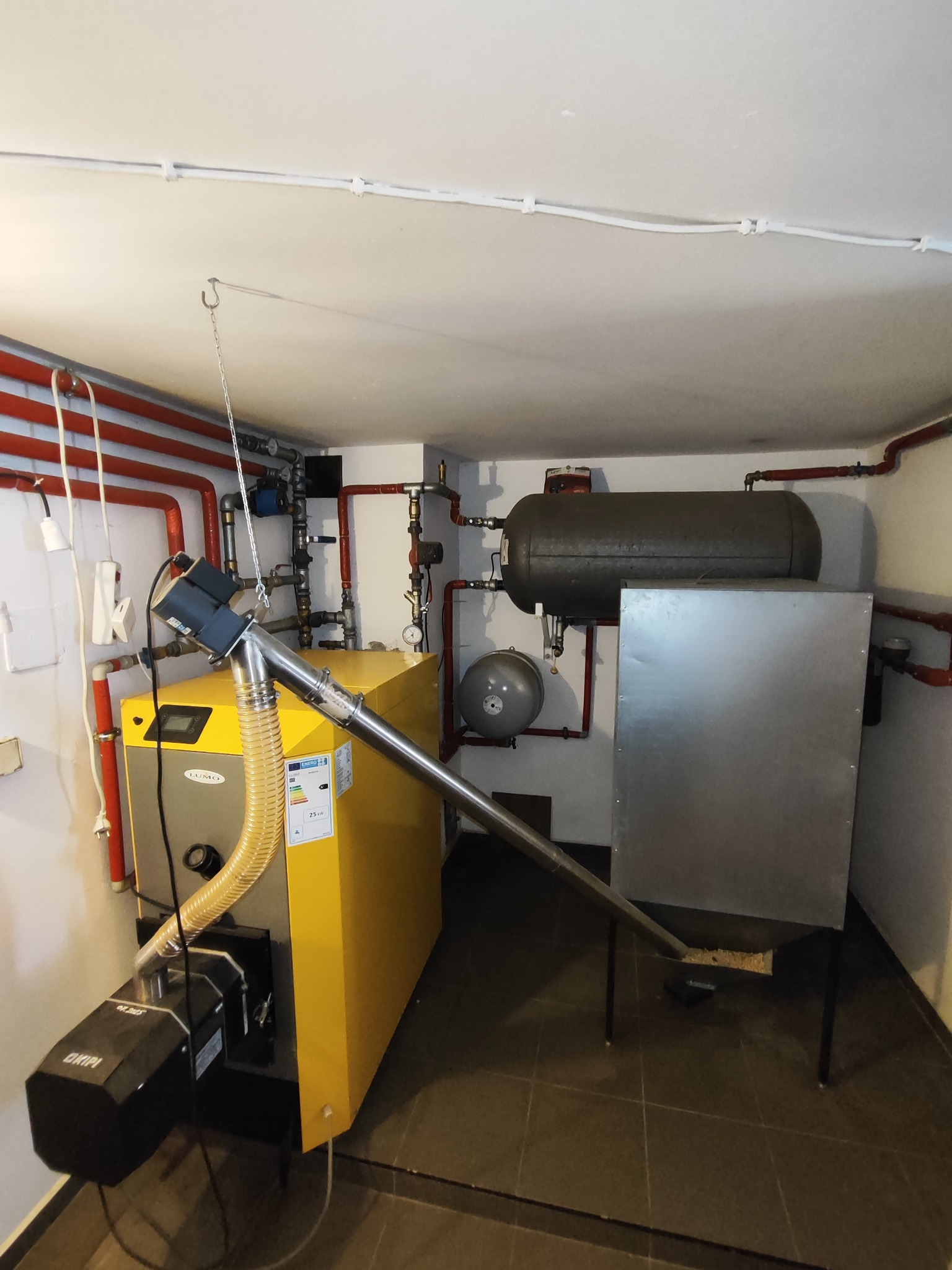 Żółty piec CO z podajnikiem ślimakowym w kotłowni, widoczne rury instalacji grzewczej i zbiornik wyrównawczy. Całość w pomieszczeniu z szarą podłogą.