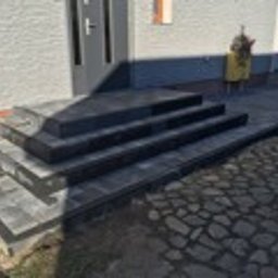 DDK BUD - Nowoczesne schody wejściowe z szarej kostki brukowej, prowadzące do drzwi domu. Ułożony chodnik z elementów kamiennych. Widoczne detale elewacji i roślinność.