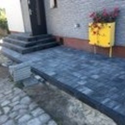 DDK BUD - Wykończony podjazd z kostki brukowej w odcieniach szarości prowadzący do schodów wejściowych. Żółta donica z kwiatami pod oknem. Kamienna ścieżka obok.