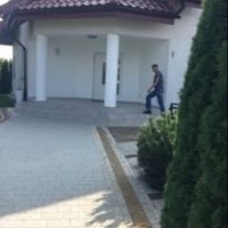 DDK BUD - Biały dom z podjazdem z jasnej kostki brukowej, oddzielonym pasem ciemniejszej kostki. Mężczyzna w oddali, krzewy i drzewa po bokach, częściowo zasłaniają widok.