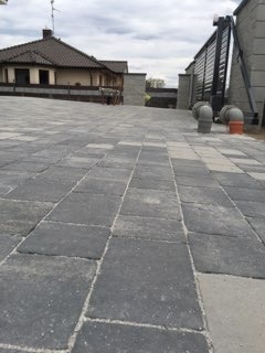 Szara kostka brukowa ułożona na posesji z widocznym domem w tle i bramą wjazdową. Widok z poziomu gruntu podkreśla regularność wzoru kostki.