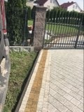 Ułożona kostka brukowa w odcieniach szarości i beżu, z widocznym obrzeżem i słupkiem bramy. Fragment samochodu z lewej strony kadru. Trawa i żywopłot w tle.