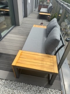 Nowoczesny taras drewniany z szarą sofą i stolikiem w Strzelcach Górnych. Deski tarasowe ułożone wzdłuż, szklana balustrada. Widok z góry, jasne oświetlenie.