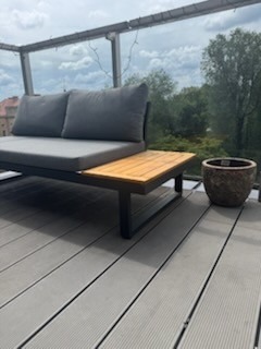 Nowoczesny taras z szarych desek kompozytowych, z sofą z szarymi poduchami i drewnianym stolikiem oraz dekoracyjną donicą. Szklana balustrada w tle.