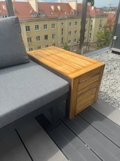 Drewniany stolik kawowy zintegrowany z sofą na balkonie, szare płytki podłogowe, widok na budynki w tle. Nietypowe rozwiązanie meblowe.