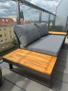 Nowoczesna sofa tarasowa z drewnianymi blatami i szarą tapicerką na balkonie z widokiem na miasto w tle. Metalowa rama, minimalistyczny design.