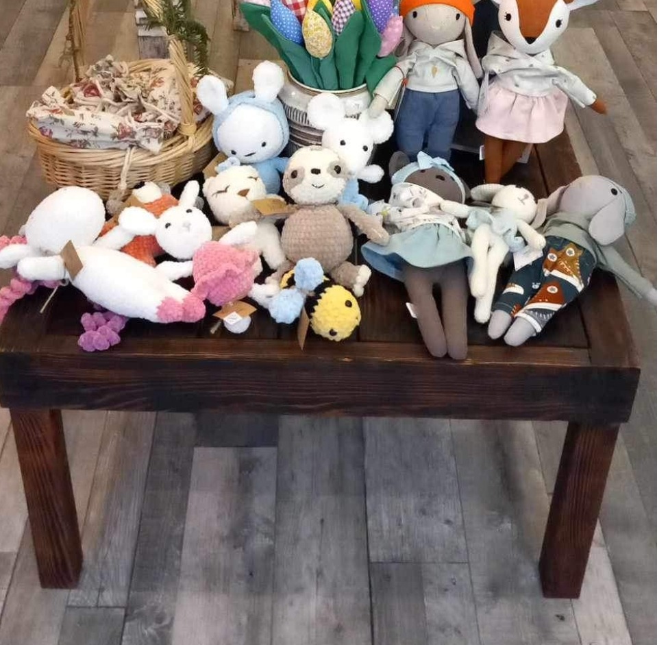 Ręcznie robione maskotki, w tym lalki, króliczki i leniwiec, ułożone na ciemnym, drewnianym stole z widoczną fakturą drewna. W tle koszyk z materiałem i bukiet tulipanów.