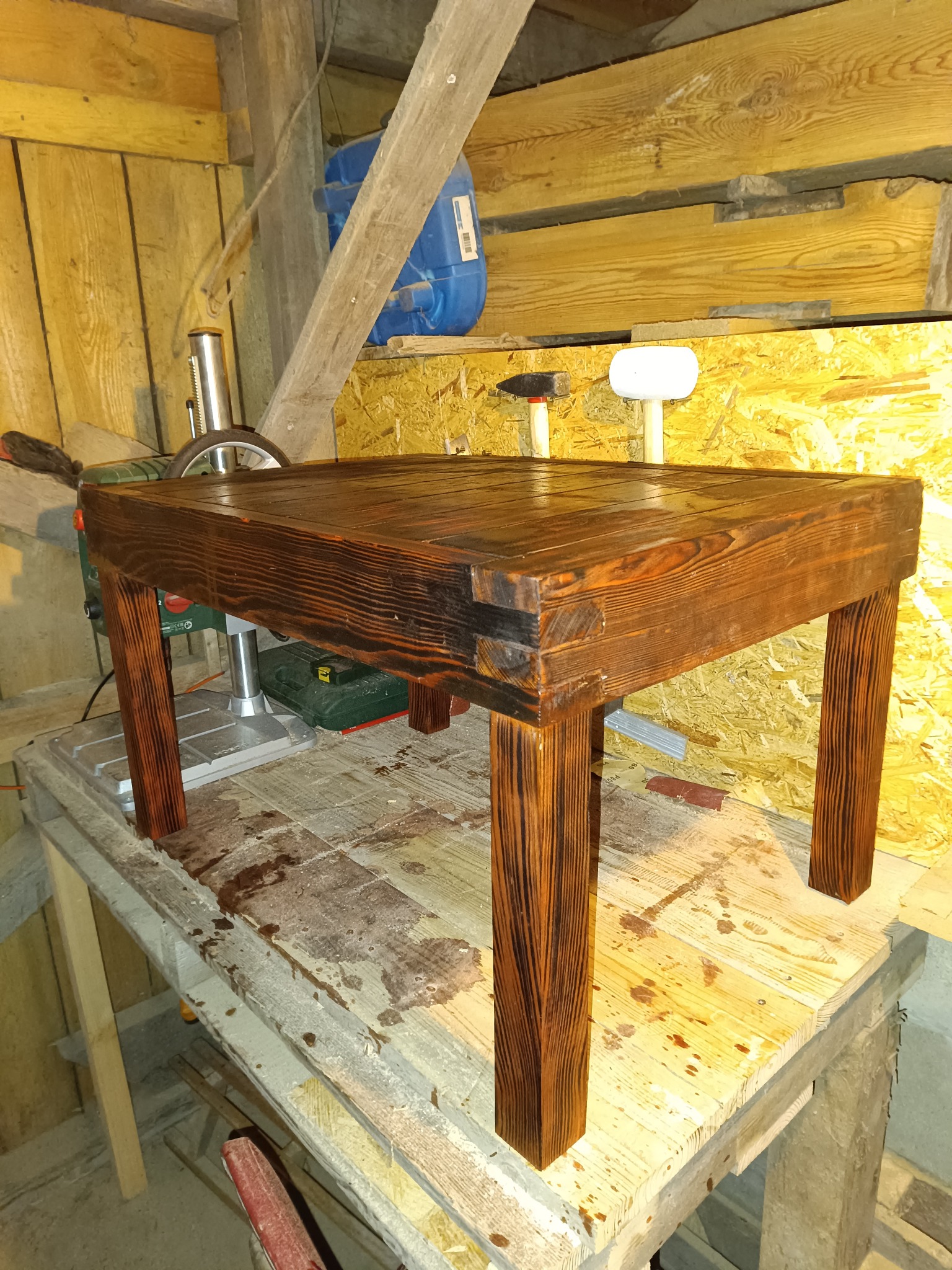 Ręcznie robiony, kwadratowy stolik kawowy z ciemnego, lakierowanego drewna, stojący na roboczym stole w warsztacie. Widoczne narzędzia i materiały stolarskie.