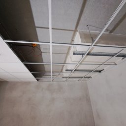 Usługi ogólnobudowlane Konrad Sobczyk - Sufit podwieszany w trakcie montażu: metalowa konstrukcja kratowa, częściowo zamontowane białe panele, widoczne instalacje. Surowa ściana w tle.
