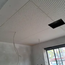 Montaż perforowanej płyty karton gips na suficie z otworem na oświetlenie, widoczne kable i surowe ściany. W tle okno z widokiem na zewnątrz.