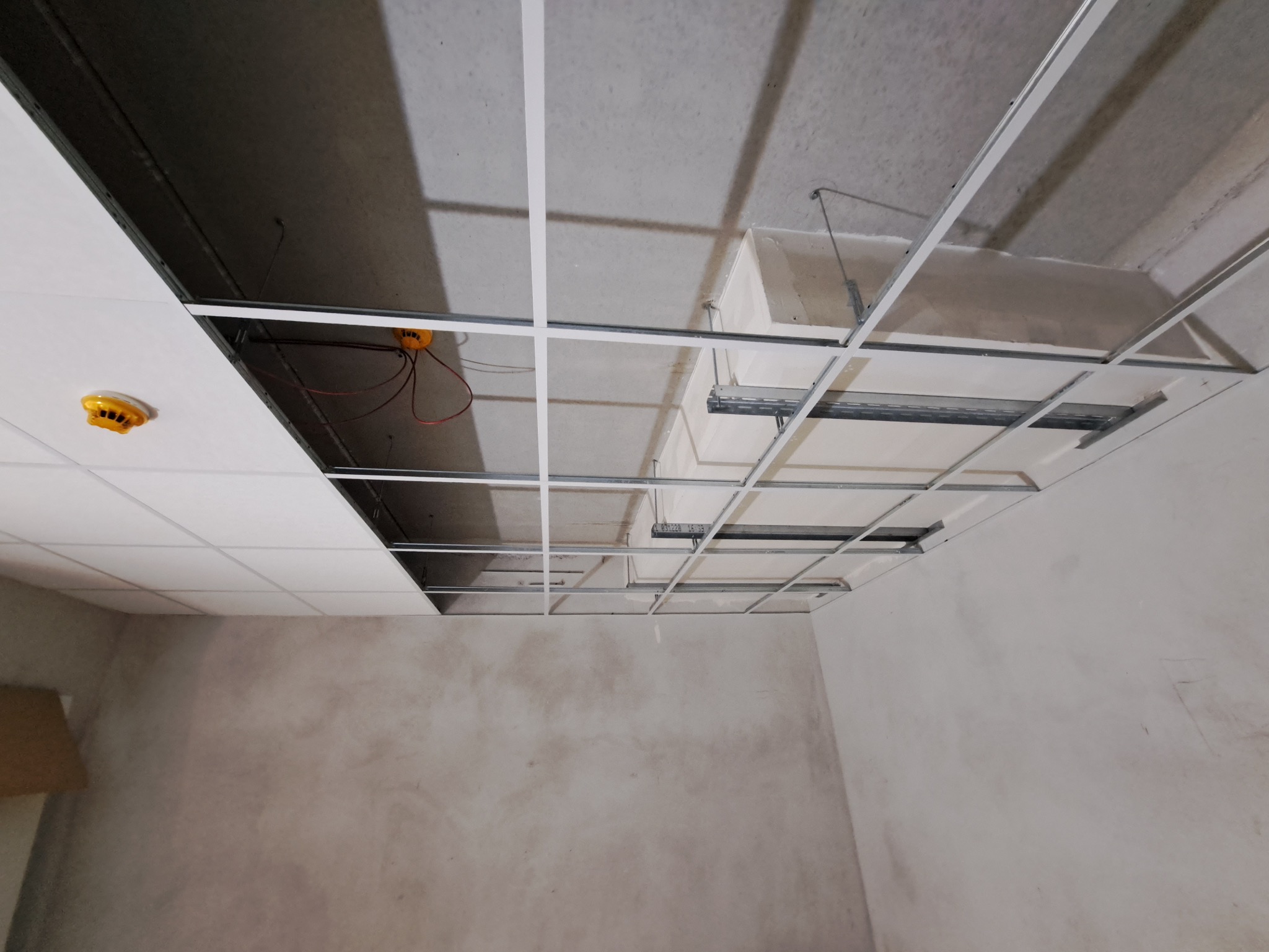 Sufit podwieszany w trakcie montażu: metalowa konstrukcja kratowa, częściowo zamontowane białe panele, widoczne instalacje. Surowa ściana w tle.