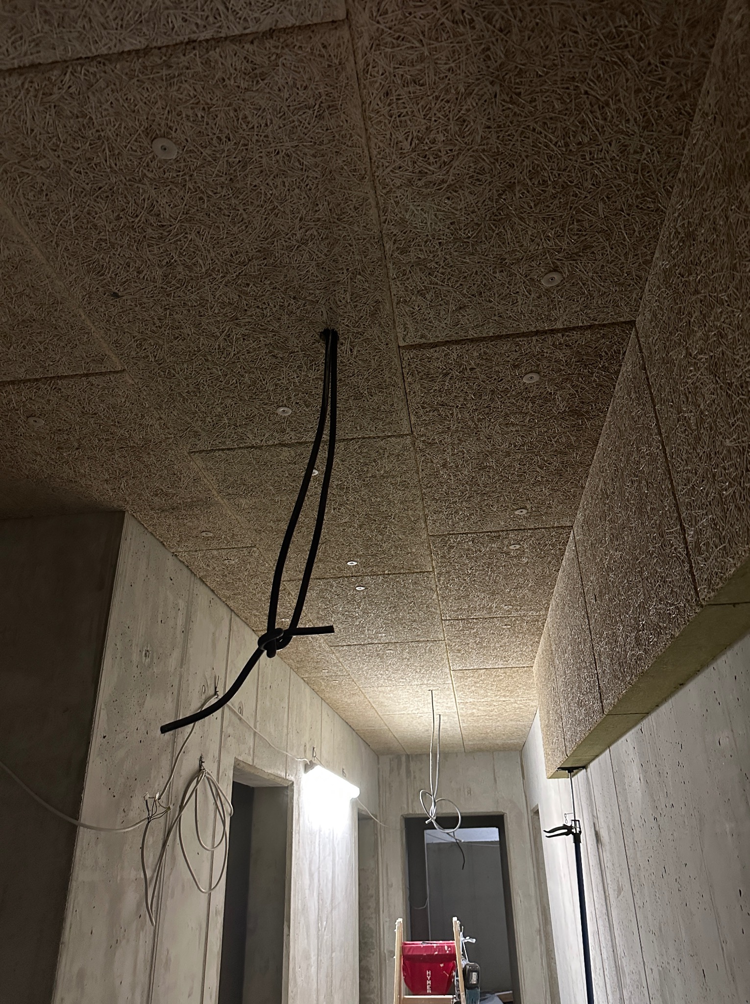 Montaż paneli akustycznych na suficie w surowym wnętrzu. Widoczne przewody elektryczne i betonowe ściany. Praca w toku, surowy stan.