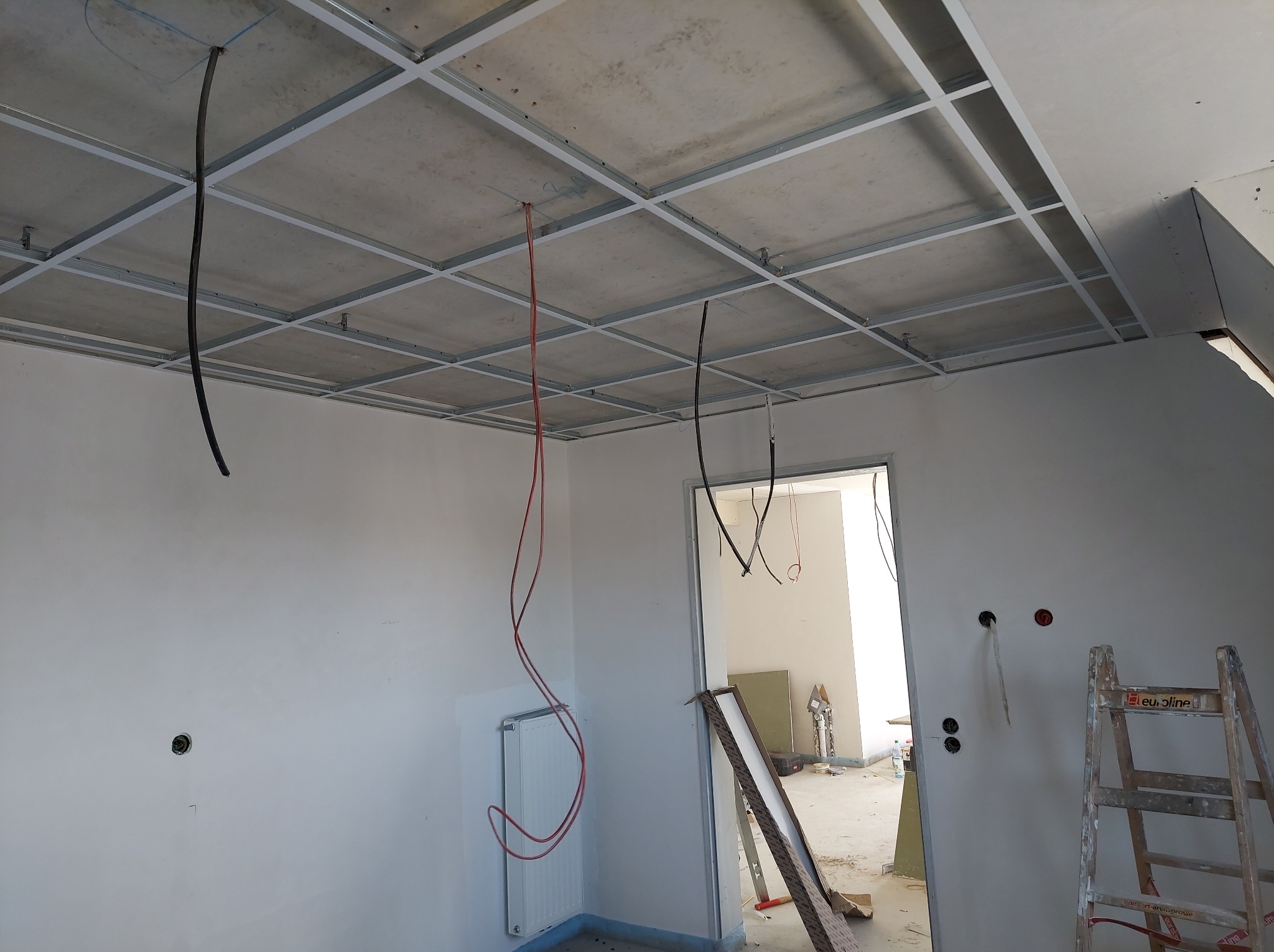 Sufit z metalową konstrukcją pod płyty, widoczne kable elektryczne, ściany przygotowane do wykończenia, drabina. Wnętrze w trakcie prac remontowych.