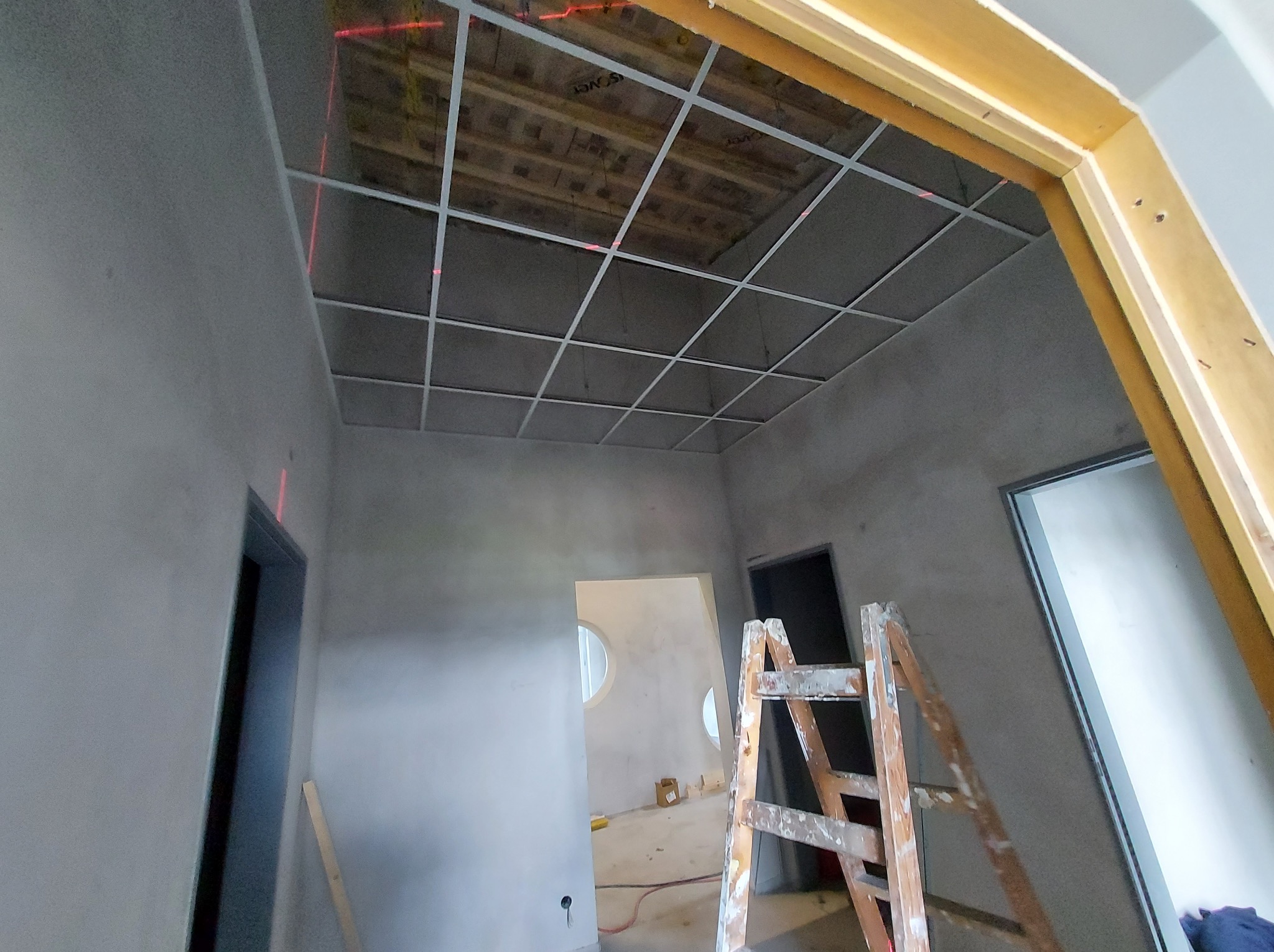 Sufit podwieszany w trakcie montażu, widoczne profile i laser krzyżowy. Wnętrze w stanie surowym, drabina malarska w pomieszczeniu. Widok na otwór drzwiowy.